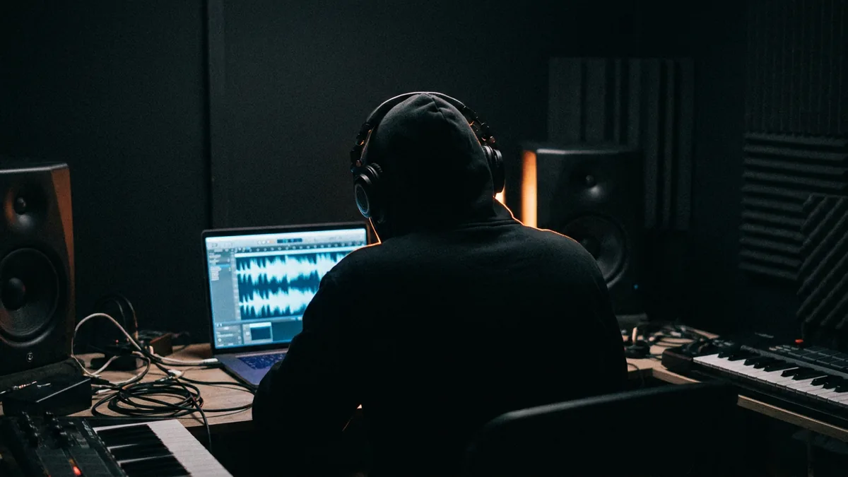 a composer alone at a home studio desk at night, headphones on, a glowing waveform on the laptop screen